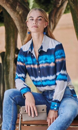Una mujer está sentada en un banco con jeans y una camisa azul tie-dye, mostrando el efecto moderno de la Camisa efecto Tie Dye.
